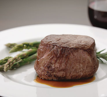 baseball_cut white plate with baseball cut of cooked beef and asparagus. Hint of red wine glass in background