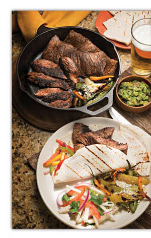 fajitas_press-release Black, cast iron skillet with cooked beef, small wooden container of guacamole, plate of grilled steak fajitas with sliced bell peppers and onions