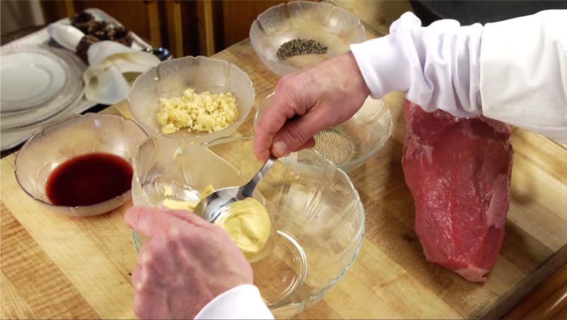 Cutting board with ingredients for preparing beef recipe, including bowl of red wine, bowl of crushed garlic, bowl of spices, large cut of beef. Chefs hands in foreground spooning mustard into large bowl