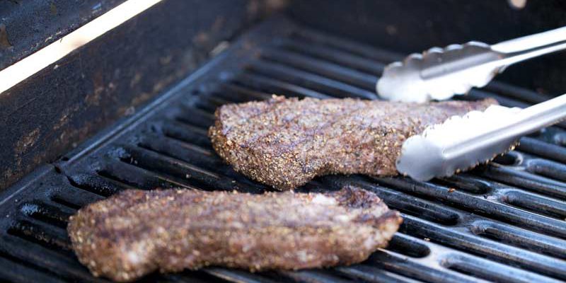 1c865a42-c8d2-4131-854d-cc9d85905ba4 Close up of 3 NY steaks on grill. Steaks look ready to come off, have criss cross grill marks. Tongs in shot reaching for steak farthest from the camera