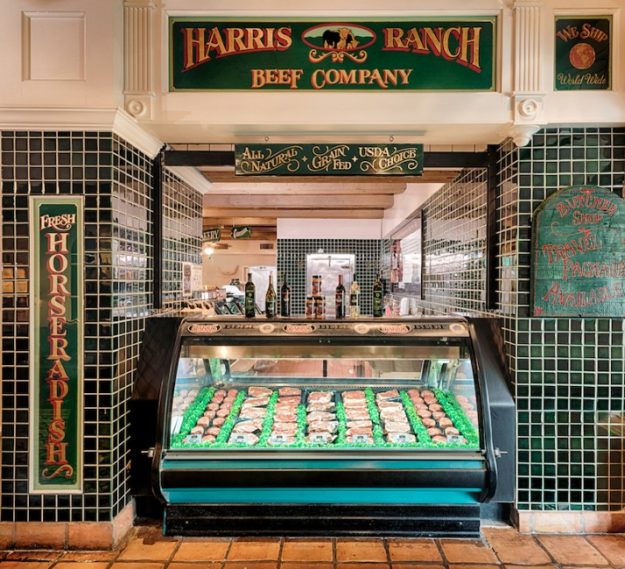 Stocked meat case at Harris Ranch Inn & Restaurant, Sign above: Harris Ranch Beef Company, All Natural, Grain Fed, USDA Choice. Green tiles on wall with white grout surround meat case. Sign on left says: Fresh Horseradish