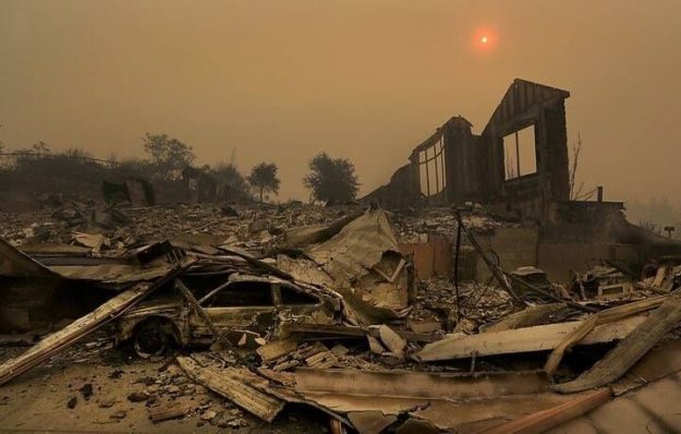 Devastation from scene of recent fire. Burned out car, collapsed structure with only one partial wall remaining, yellow sun high in smokey sky, shrouded with orange ring